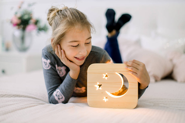 Little pretty 10 years old girl, lying on the bed at cozy light bedroom at home, and posing to camera with wooden stylish night lamp with cut out moon and stars picture. Wooden decorations - Photo, Image