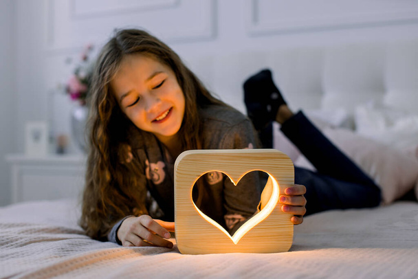 Children, emotions and night lamps concept. Indoor horizontal shot of beautiful child school girl, lying on the bed at home and having fun with stylish hand made wooden night lamp with heart picture - Fotó, kép