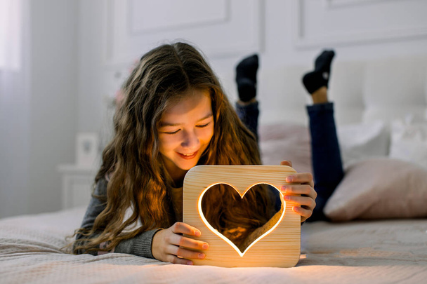 Children, emotions and night lamps concept. Indoor horizontal shot of beautiful child school girl, lying on the bed at home and having fun with stylish hand made wooden night lamp with heart picture - Foto, immagini