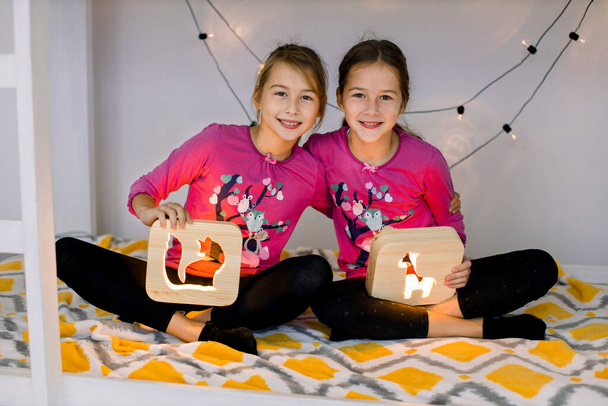 Happy children, two smiling cute 10 year old girls sisters, in childs room on a bunk-bed, sitting in lotus position and holding wooden night lamps with cut out pictures. - Photo, Image