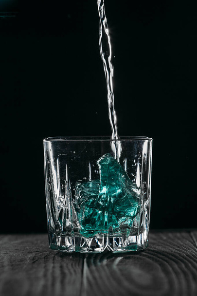 Glass with blue ice cubes. Pouring gin into a glass on a wooden table. Gin in a glass on a black background. - Photo, image