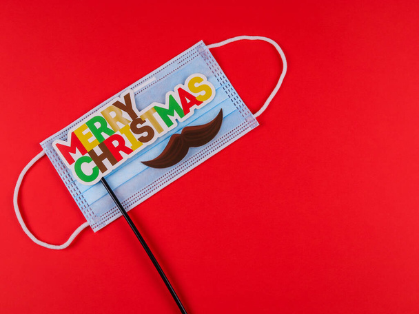 Máscara e inscrição em uma vara: Feliz Natal jazem à esquerda em um fundo vermelho com espaço para texto à direita, vista superior close-up. - Foto, Imagem