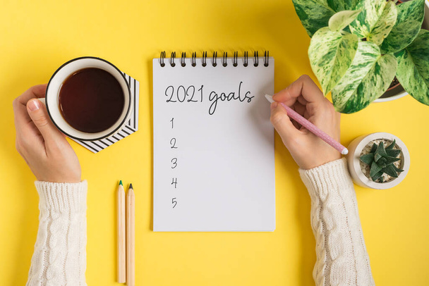 Desktop of woman hands with blank notepad writing 2021 goals text . Flat lay of yellow working table background with cup of tea and plants. Top view mock up  - Photo, Image
