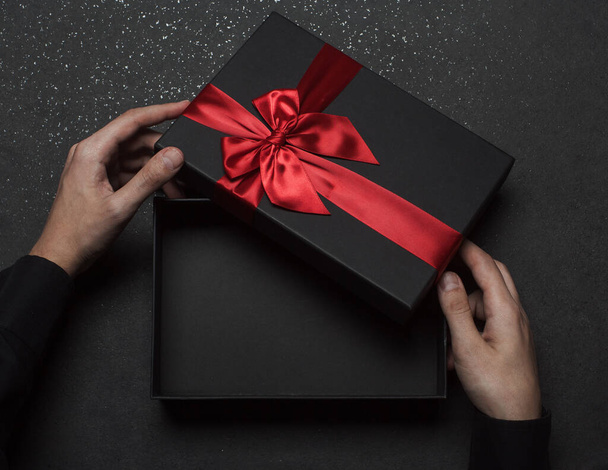 The hands of a guy in a black shirt open a gift box with a large red bow. Empty box. Granite background. - 写真・画像