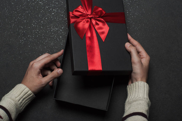 Hands in a sweater opening a black gift box with a red bow on a black granite background. - 写真・画像