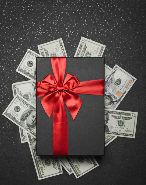 Μαύρο κουτί δώρου με κόκκινη κορδέλα και ένα μεγάλο φιόγκο σε ένα σωρό χρήματα. Δώρο σε μια επιφάνεια γρανίτη. - Φωτογραφία, εικόνα