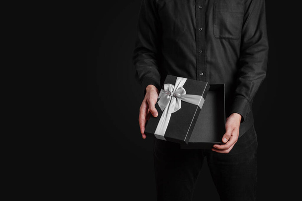 Ένας τύπος με μαύρο πουκάμισο κρατάει ένα ανοιχτό κουτί δώρου με ασημένιο φιόγκο. Δώρο στο χέρι. Μαύρο φόντο. - Φωτογραφία, εικόνα
