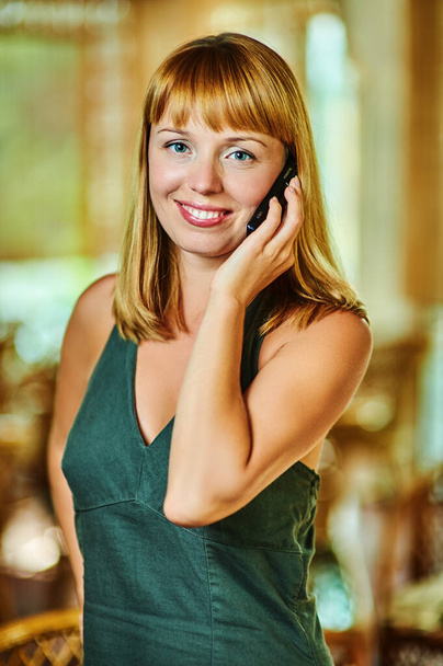 Молодая счастливая рыжая женщина в зеленом платье разговаривает по мобильному телефону на фоне большого зала с плетеными стульями. - Фото, изображение