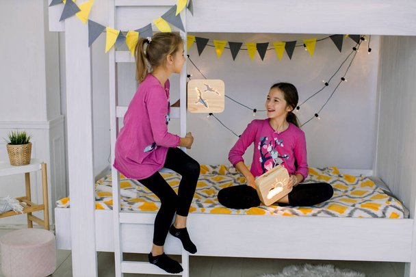 Дети играют в спальне. Две смешные счастливые девочки 10 лет, сестры в красочных пижамах, веселятся на двухъярусной кровати, держат деревянные ночные лампы - Фото, изображение