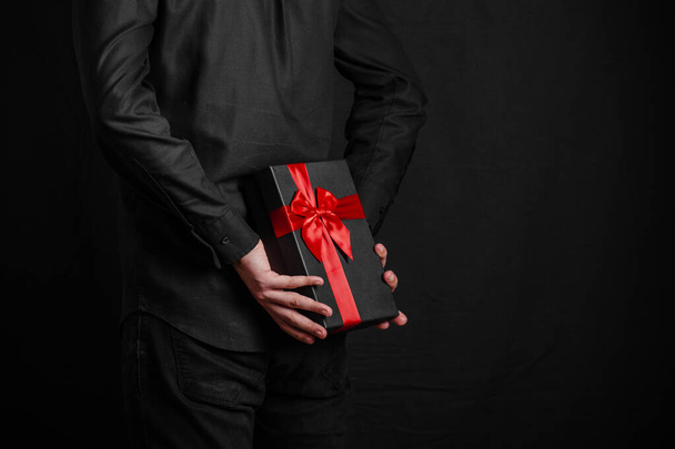 Ο άντρας με το μαύρο πουκάμισο κρατάει ένα δώρο με μια κόκκινη κορδέλα πίσω από την πλάτη του. Μαύρο κουτί δώρου στα χέρια. Έκπληξη. Θέση για επιγραφή - Φωτογραφία, εικόνα
