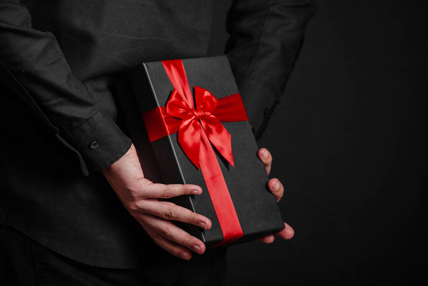 Ο άντρας με το μαύρο πουκάμισο κρατάει ένα δώρο με μια κόκκινη κορδέλα πίσω από την πλάτη του. Μαύρο κουτί δώρου στα χέρια. Έκπληξη. Θέση για επιγραφή - Φωτογραφία, εικόνα