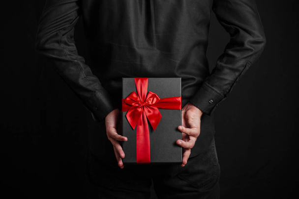 Man in een zwart shirt heeft een geschenk met een rood lint achter zijn rug. Zwarte geschenkdoos in handen. Verrassing. - Foto, afbeelding