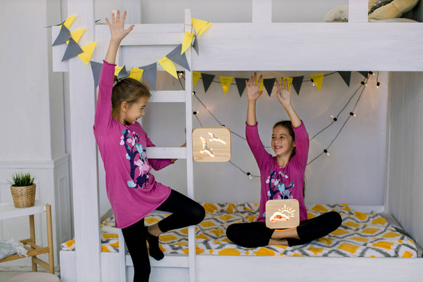 Children playing in the bedroom. Two funny happy girls 10 years old, sisters in colorful pyjamas, having fun on a bunk bed, holding wooden night lamps - Фото, изображение
