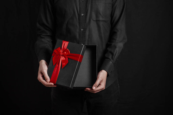 Un tipo con una camisa negra sobre fondo negro sostiene una caja de regalo abierta con un lazo rojo. - Foto, imagen