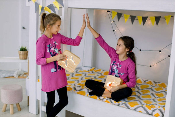 Children playing in the bedroom. Two funny happy girls 10 years old, sisters in colorful pyjamas, having fun on a bunk bed, holding wooden night lamps - Photo, image