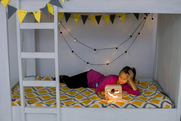 The cute girl is ready to sleep in the bed. Evening night time. Little girl lying on stylish decorated bunk bed and looking at the wooden night lamp - Φωτογραφία, εικόνα