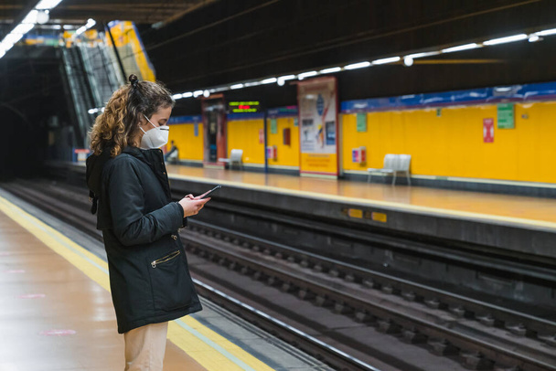 Uusi normaali. Nuori tyttö käyttää älypuhelinta metrossa. Teini-ikäinen käyttää älypuhelintaan metrossa. Teini-ikäinen naamiossa. Nuori tyttö, jolla on suojaava naamio. Pandemia. - Valokuva, kuva