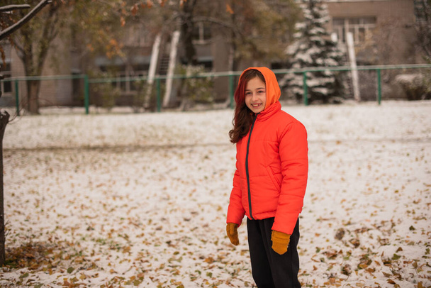 ένα κορίτσι με ένα φωτεινό πορτοκαλί ζεστό σακάκι. Κορίτσι με πορτοκαλί μπουφάν με χιονισμένο καιρό. Έφηβη κοπέλα το χειμώνα - Φωτογραφία, εικόνα