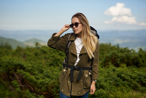 Stylish girl traveller in sunglasses is standing at the hight pick of the mountain . Hiker is looking to the right side of herself . Concept of tourist at the head of big mountain with modern wear and a backpack - Photo, Image