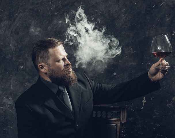 Stevige zelfverzekerde man met baard in pak met glas whisky en rook - Foto, afbeelding