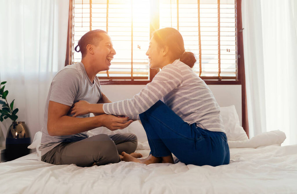Portrait of happy young adult Asian couple tickling each other on bed together in bedroom interior scene. 30s candid mature husband and wife smiling. Marriage and happy relationship life concept - Photo, Image