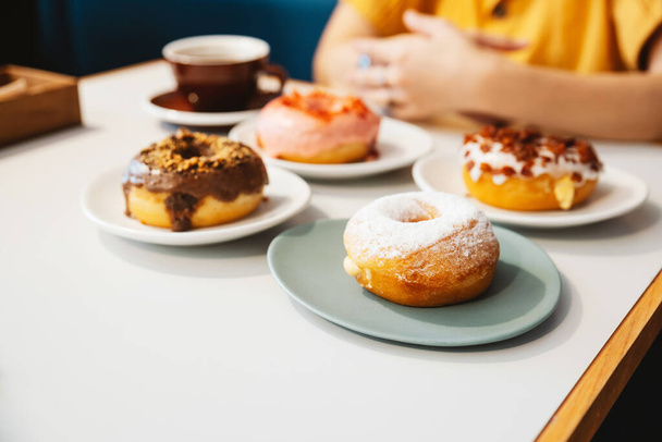 Donuts on the table include Strawberry pink, chocolate, sugar glazed and bacon, cheese donut on the table with the woman wearing yellow shirt in background in modern cafe. Enjoyment female lifestyle. - Photo, Image