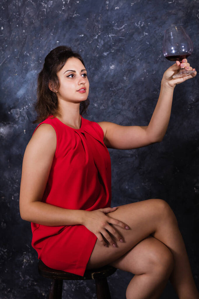Gyönyörű fiatal hölgy piros ruhában ül egy pohár borral. - Fotó, kép