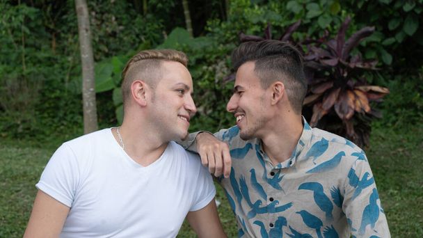 Couple gay profiter d'une journée dans le parc, l'un d'eux a son bras reposant sur l'épaule de l'autre, les deux regarder l'autre et sourire. - Photo, image
