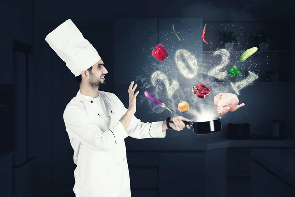 Happy male magician chef cooking on a saucepan with flying food ingredients and smoke shaped of numbers 2021 in the kitchen - Photo, image