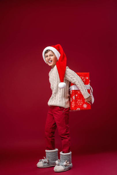 Happy laughing boy in Santa hat with Christmas gift on red background. the child is happy and holds a big gift in his hands. New year sales, advertising, discounts. Copy space. - Photo, Image