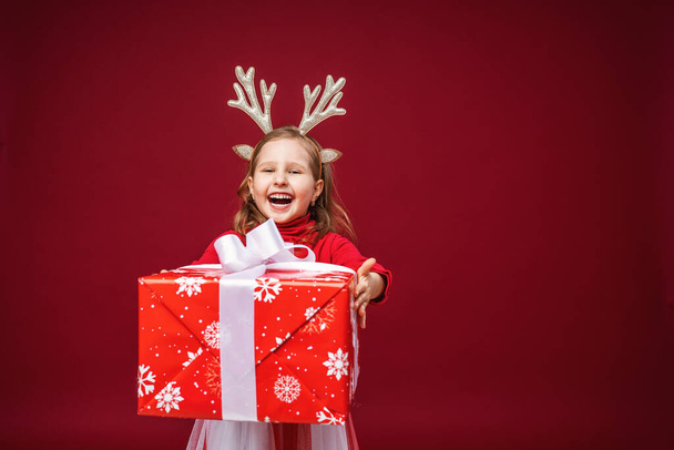 Happy little girl 4 years old in reindeer antlers on a bright red background. child is happy and holds gift in his hands and stretches it forward. Give gifts. New year sales, advertising, discounts. - Φωτογραφία, εικόνα