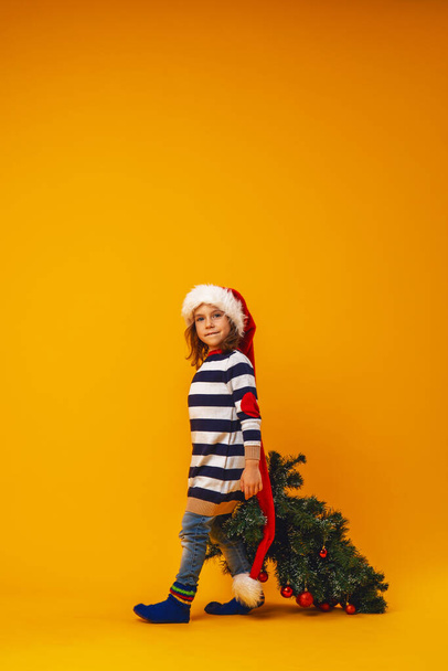 happy little girl in a striped sweater, Santa hat on her head, with a Christmas tree decorated with red balloons, walks on a yellow background. Copy space. - Valokuva, kuva