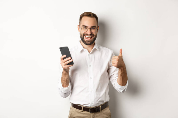 Tyytyväinen liikemies osoittaa peukalot ylös käytön jälkeen matkapuhelin, seisoo tyytyväinen valkoisella taustalla - Valokuva, kuva