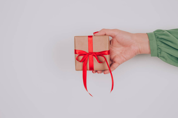 Γυναικείο χέρι κρατώντας το παρόν κουτί διακοσμημένα με κόκκινο τόξο. Χριστουγεννιάτικες διακοπές, Πρωτοχρονιά ή εορταστική ιδέα γενεθλίων. Προετοιμασία οικογενειακών δώρων για τα Χριστούγεννα - Φωτογραφία, εικόνα