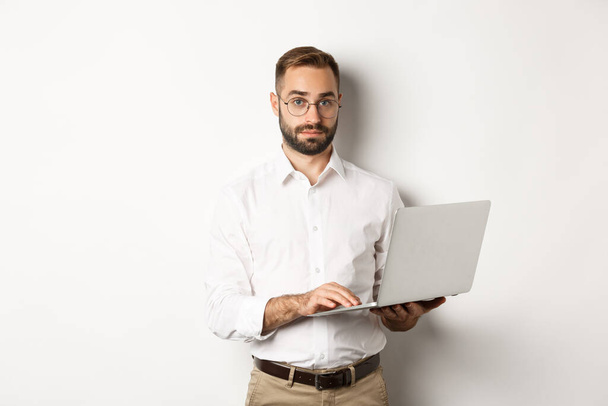 Για δουλειές. Νέος όμορφος επιχειρηματίας που εργάζεται σε φορητό υπολογιστή, κάνει τη δουλειά του στον υπολογιστή, στέκεται πάνω από λευκό φόντο - Φωτογραφία, εικόνα