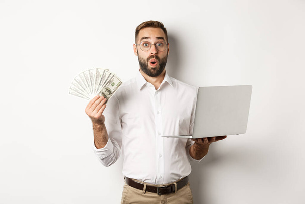 Affari ed e-commerce. Uomo guardando stupito con i soldi di reddito, lavorando online, utilizzando il computer portatile, in piedi su sfondo bianco - Foto, immagini