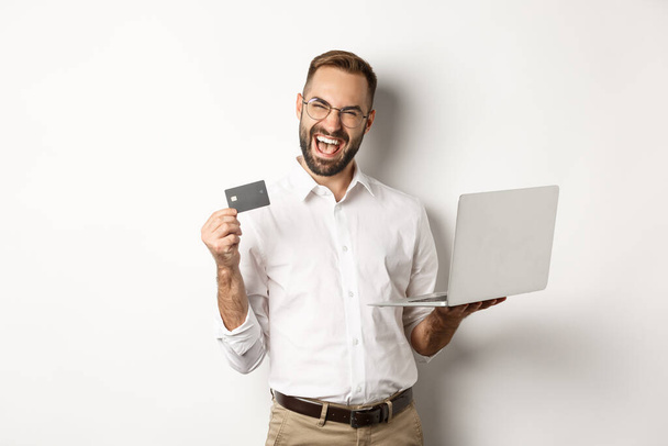 Verkkokaupoissa. Komea mies näyttää luottokorttia ja käyttää kannettavaa tietokonetta tilauksesta internetissä, seisoo valkoisella taustalla - Valokuva, kuva