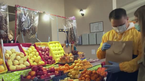 Junges Paar mit Schutzmaske und Handschuhen wählt eine frische Mandarine, steckt sie in eine Papiertüte und gibt sie dem Verkäufer auf dem Markt - Filmmaterial, Video