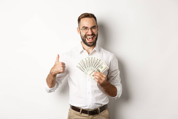 Ενθουσιασμένος πλούσιος που κρατάει χρήματα, επιδεικνύοντας τον αντίχειρά του σε επιδοκιμασία, στέκεται πάνω από λευκό φόντο - Φωτογραφία, εικόνα