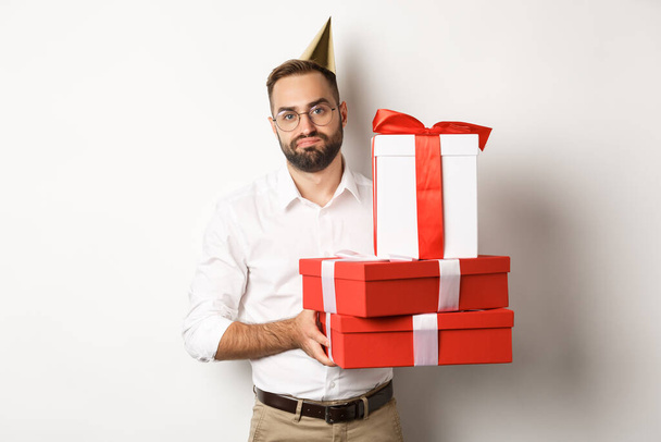 Holidays and celebration. Displeased guy holding birthday gifts and looking disappointed, dislike presents, standing over white background - 写真・画像