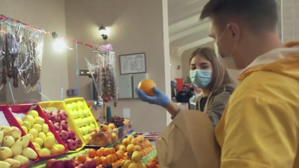 Pareja joven con máscara protectora y guantes elige una naranjas frescas, las pone en una bolsa de papel ang se lo da al vendedor en el mercado - Metraje, vídeo