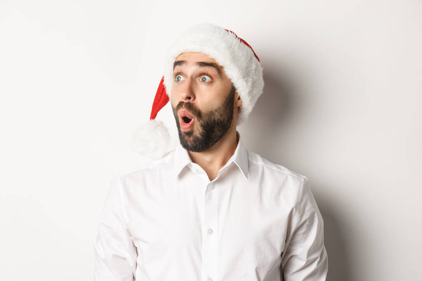 サンタの帽子の驚くべき髭の男のクローズアップ,驚きの顔で左を見て,クリスマスプロモーションの提供,白い背景の上に立って - 写真・画像