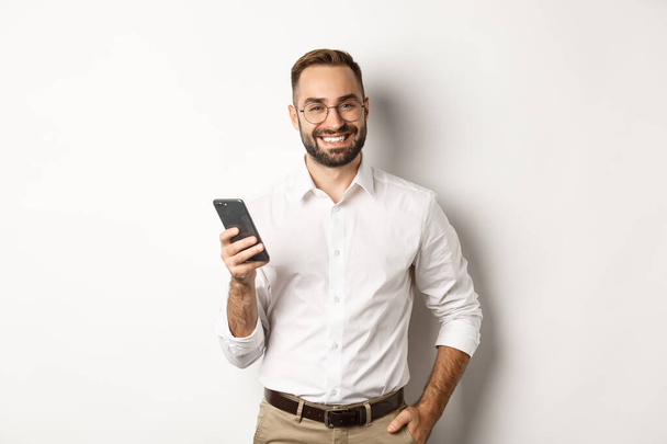 Fešák manažer pomocí chytrého telefonu a s úsměvem potěšen, posílání SMS, stojící nad bílým pozadím - Fotografie, Obrázek