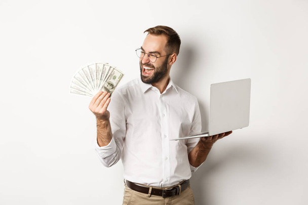 Affari ed e-commerce. Uomo d'affari di successo utilizzando il computer portatile per lavoro e tenendo i soldi, in piedi su sfondo bianco - Foto, immagini