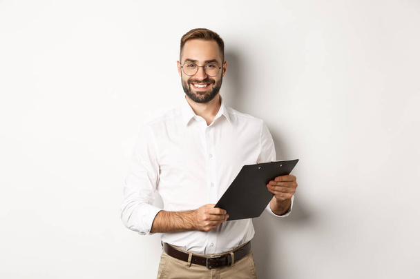 Handsome manager in glasses working, using clipboard, standing over white background - Photo, Image