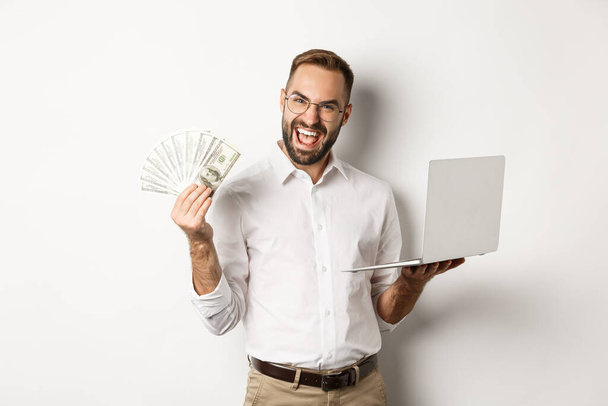 Бизнес и электронная коммерция. Возбужденный бизнесмен с деньгами и ноутбуком, работающий в сети, стоящий на белом фоне - Фото, изображение