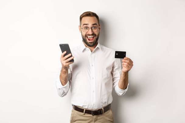 Business and online payment. Excited man paying with mobile phone and credit card, smiling amazed, standing over white background - Foto, Imagen