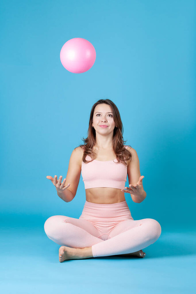 a young woman in a pink sports top and leggings sits cross-legged and throws a ball up, isolated on a blue background - 写真・画像