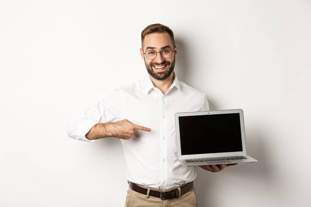 Professional manager showing webpage on laptop screen, pointing at computer, standing over white background - Fotoğraf, Görsel