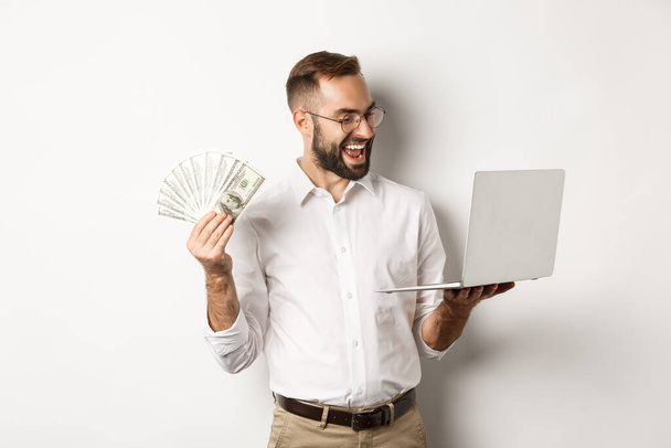 Бизнес и электронная коммерция. Довольный бизнесмен делает работу на ноутбуке и держит деньги, улыбаясь счастливым, стоя на белом фоне - Фото, изображение